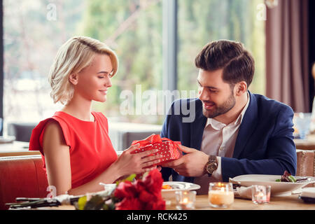 Young man and woman on date in restaurant sitting at table guy giving red gift box to lady smiling happy surprised Stock Photo
