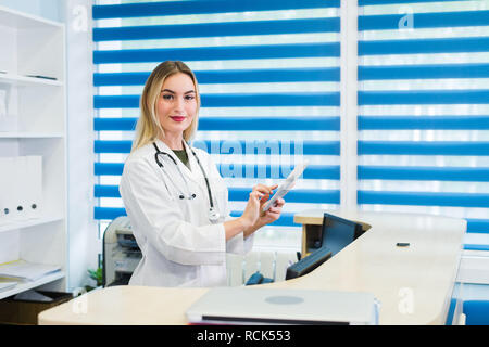 Smiling female doctor wearing a scrub and working at the hospital reception, she is writing a medical report on a tablet computer and scheduling appointments Stock Photo