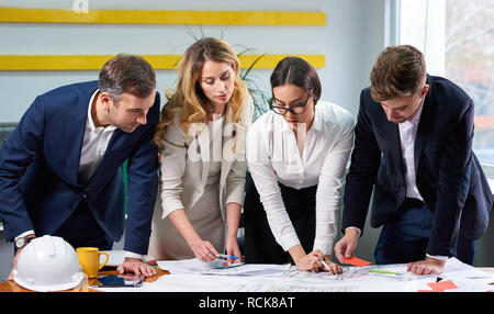 Team of mature women and men at meeting table discussing a construction plan blueprint with hard hat in office Stock Photo