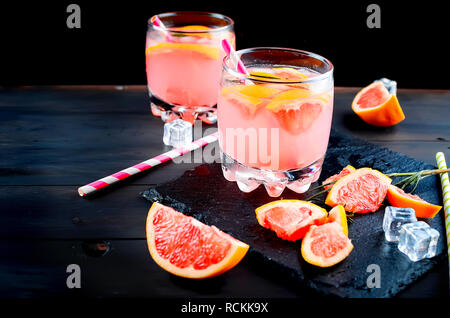 grapefruit juice or cocktail in glasses and pieces of grapefruit, ice cubes, straws on a dark background Stock Photo