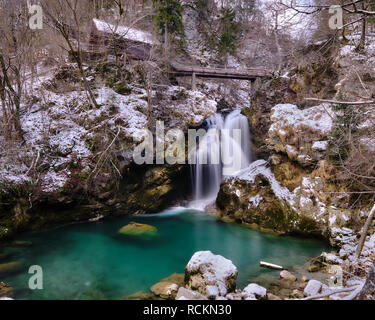 Cabin in Vintgar gorge in winter. Waterfall dropping from under a wooden bridge into a green pool of water. In a rocky forest after a dusting of snow Stock Photo