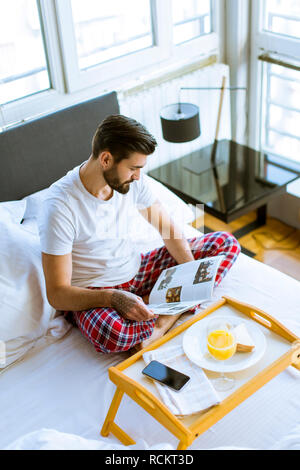 Young bearded  man having breakfast in bed and reading magazine in the room Stock Photo