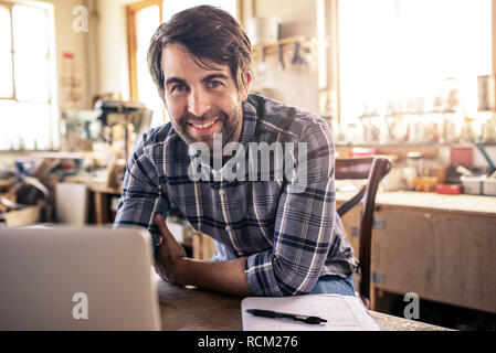 Woodworker smiling while using a laptop in his workshop Stock Photo
