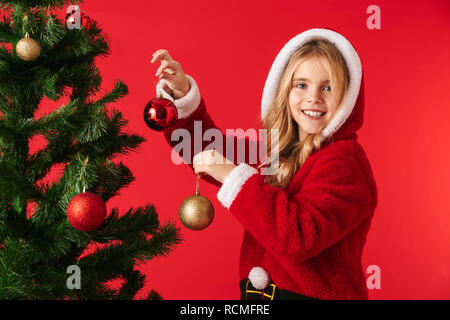 Cheerful little girl wearing Christmas costume standing isolated over red background, decorating Christmas tree Stock Photo