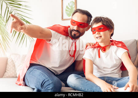 Father showing little son view out the window smiling dreamful wearing superheroe costumes together at home sitting on sofa Stock Photo