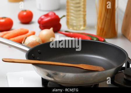 An empty pan with a wooden spatula for stirring stands on a gas stove, next to fresh vegetables and pasta Stock Photo