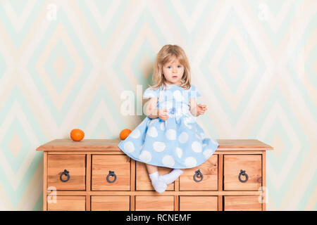 A little baby girl in her room sitting cross-legged on chest of drawers with tangerines on the rhomb wallpaper background. Child in blue polka dot dress and white socks attentively looking at camera. Stock Photo