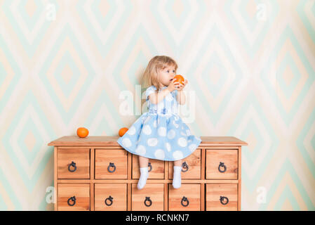 Portrait of little girl in blue polka dot dress and white socks with tangerine in hands sitting on wooden commode and going to eat it. Wallpaper on the background. Vitamins, fruit and children's joy Stock Photo