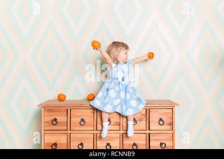 Little girl holding tangerines or oranges in her hands while sitting on the wooden wardrobe. Wearing light blue dress. Christmas holidays is over. Happy smiling baby raising hands up looking aside Stock Photo