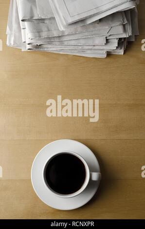 Overhead shot of a cup and saucer of black coffee placed on a light wood laminate table with a pile of newspapers. Stock Photo