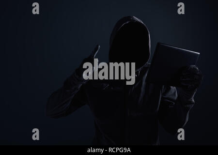 Hooded computer hacker with obscured face using digital tablet in cybercrime and cybersecurity concept, low key with selective focus Stock Photo