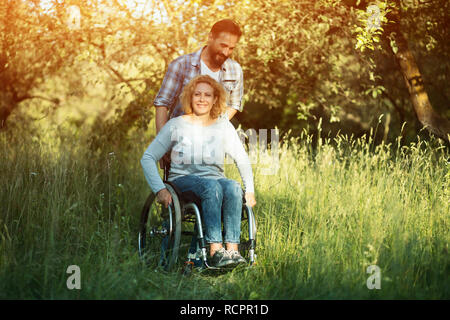 Smiling woman in wheelchair with husband in the park on sunny day Stock Photo