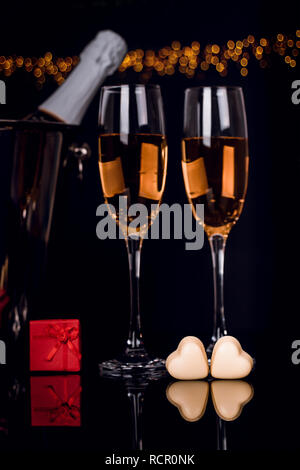 Two wine glasses, gift in a box and two heart-shaped chocolates on black background with golden lights . Love, Valentines day concept Stock Photo