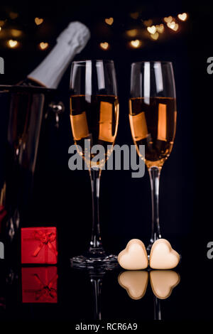Two wine glasses, gift in a box and two heart-shaped chocolates on black background with lights. Love, Valentines day concept Stock Photo
