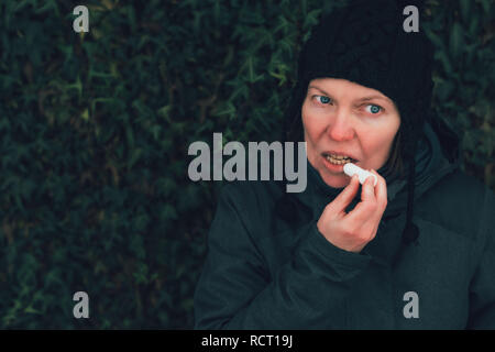 Woman applying lip balm out on the street on a cold winter day against the common ivy wall Stock Photo