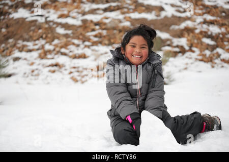 Smiling Latino girl sitting on snow covered mountain, making a snowman and wearing winter clothes. Stock Photo