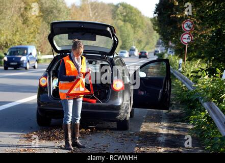 Car breakdown, female driver on the hard shoulder of a country road, wearing a reflective vest, putting up a warning triangle Stock Photo