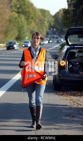 Car breakdown, female driver on the hard shoulder of a country road, wearing a reflective vest, putting up a warning triangle Stock Photo