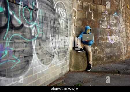 Man wearing a mask is surfing on a laptop computer in front of graffiti covered wall, symbolic image for computer hacking Stock Photo