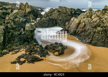 Slow shutter speed was used to capture this image of a wave swirling though the rocks on the beach.  The receding water created curves of white foam Stock Photo