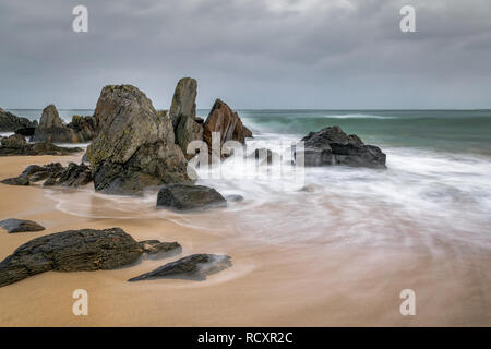 Blurred waves swirling around rocks on a sandy beach in Ireland.  The sky is stormy but the blurred waves create a soft light around the rocks. Stock Photo