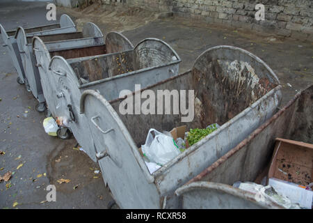 Old green dumpsters on the street. Household waste in the city. Old broken silver garbage container. Item garbage collection near the doors of industrial building - Image Stock Photo