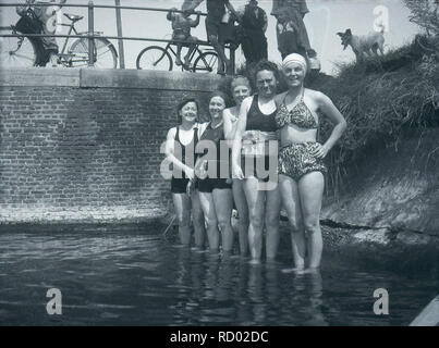 circa 1940, historical, ladies in bathing costumes of the day standing in a shallow part of a river at the side, ready for a swim or bath, with on lookers standing above on the riverbank, England, UK. One of the ladies is wearing what looks like a lifebelt around her waist. Stock Photo