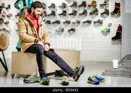 Man in winter jacket trying shoes for mountain hiking sitting in the fitting room of the modern sports shop Stock Photo