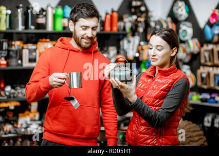 Young couple dressed in red sportswear choosing dishes for camping in the shop with travel equipment Stock Photo