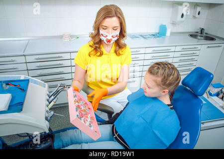 Female pediatric dentist medic making a treatment for adorable little smiling girl sitting in chair in a hospital. Dentist and child in cabinet. Littl Stock Photo