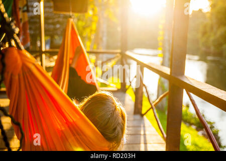 Woman lying in a hammock on the porch of a cabine, enjoying the sunset over a river in Upper Suriname Stock Photo
