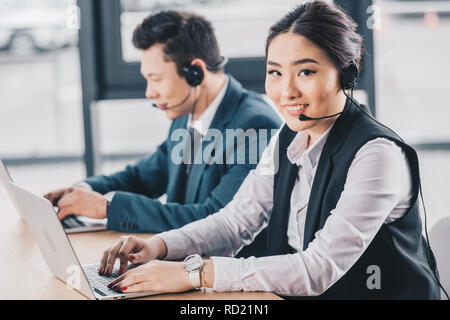young woman in headset smiling at camera while working with male colleague in call center Stock Photo