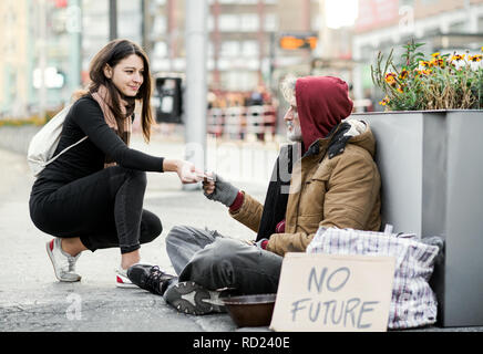 Young woman giving money to homeless beggar man sitting in city. Stock Photo