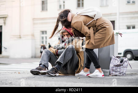 Young woman giving money to homeless beggar man sitting in city. Stock Photo