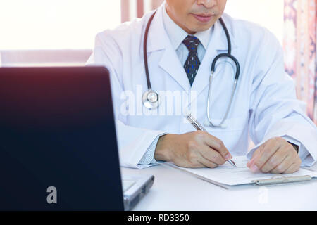 Male doctor with stethoscope writing down on a patient record in front of laptop computer notebook in a hospital office. Side copy space included. Stock Photo