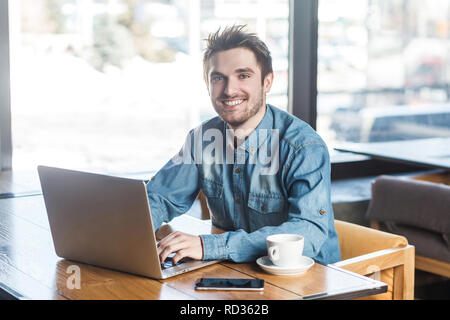 Portrait of handsome cheerful bearded young businessman in blue jeans shirt are sitting in cafe and typing message on laptop with toothy smile and loo Stock Photo