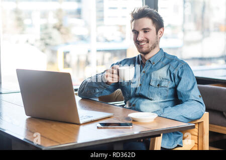 Portrait of handsome happy bearded young businessman in blue jeans shirt are sitting in cafe and have a break with cup of coffee and toothy smile are  Stock Photo