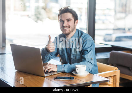 Everything alright! Portrait of handsome satisfied bearded young freelancer in blue jeans shirt are sitting in cafe and working on laptop with toothy  Stock Photo