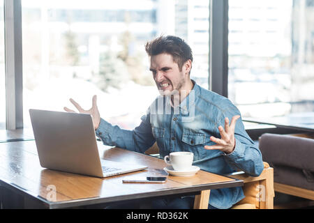 Portrait of aggressive unhappy young businessman in blue jeans shirt are sitting in cafe and having bad mood are admonishing a worker through a webcam Stock Photo