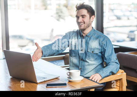 Welcome! Portrait of satisfied happy young businessman in blue jeans shirt are sitting in cafe and greeting a worker through a webcam and showing hand Stock Photo