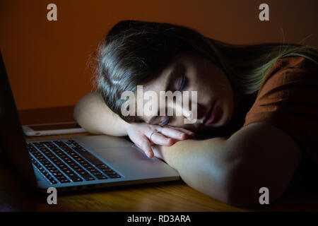 A teenage girl asleep with her head resting on the desk in front of her laptop. Stock Photo