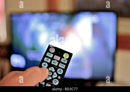 Closeup view of a television remote in hand with the tv blurred in the background. Stock Photo