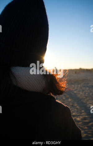 detail of back of womans head with hat and shoulders with her auburn coloured hair lit up by sun rays whilst walking on a beach at dusk in winter Stock Photo