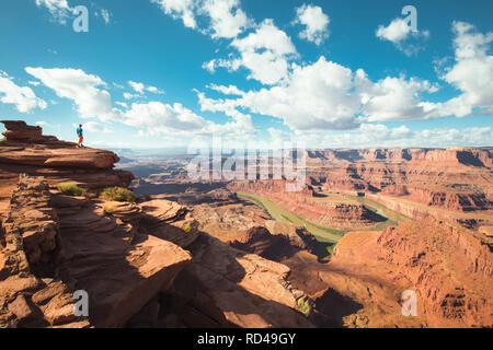 A young male hiker is standing on the edge of a cliff enjoying a dramatic overlook of the famous Colorado River, Dead Horse Point State Park. Utah, US Stock Photo