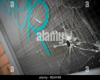 Bullet hole and cracks in a reinforced window with gang graffiti Stock Photo