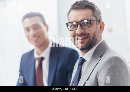 handsome bearded businessman in eyeglasses smiling at camera, young colleague standing behind Stock Photo