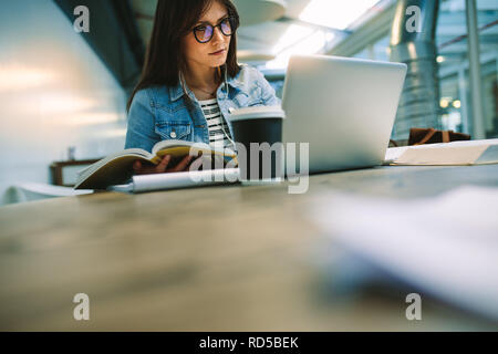 Girl with a book looking at laptop while sitting in college campus. Concentrated female student studying at university canteen. Stock Photo