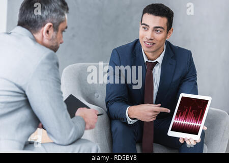 smiling young businessman pointing at digital tablet with graphs on screen and looking at male colleague during conversation Stock Photo