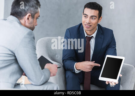 smiling young businessman pointing at digital tablet with blank screen and looking at male colleague during conversation Stock Photo