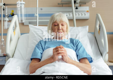 senior woman lying in bed with folded hands and praying in hospital Stock Photo